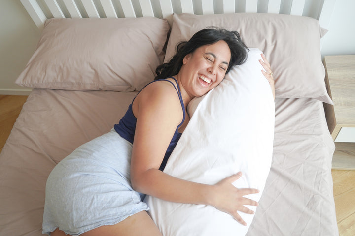 Woman laughing with eyes closed lying on a bed cuddling a Kind Face Wool Body Pillow pregnancy pillow